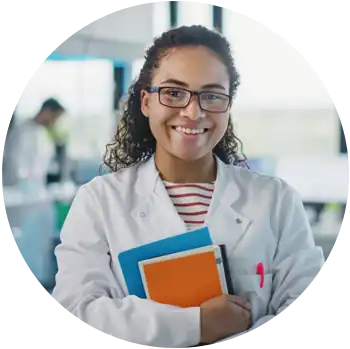A young tan-skinned female scientist in a white lab coat holds books while smiling at the camera.