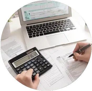 A person with light-skinned hands uses a calculator and fills out tax forms in front of a laptop.